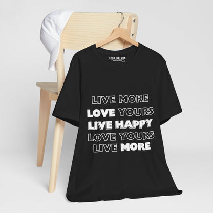 a black 100% cotton Dear No One Clothing shirt saying Live More, Love Yours, Live Happy in white lettering. the shirt is on a wooden hanger which hangs on a brown wooden chair. A white hat is also hanging on the chair.